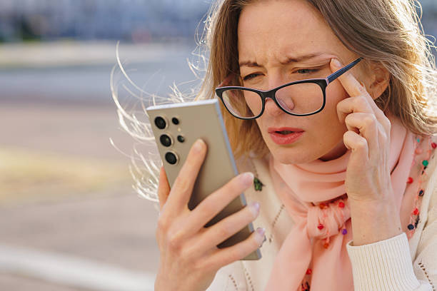 istock Frustrated woman squinting at smartphone screen while wearing glasses, symbolizing vision problems, digital eye strain, and poor eyesight in bright outdoor conditions. 2219805666