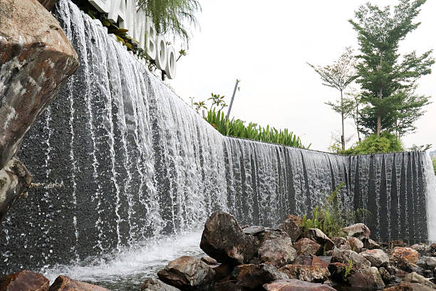 A wide, impressive artificial waterfall cascades down a dark wall into a rocky pool at its base, surrounded by green foliage and a clear sky, with a partial view of a \