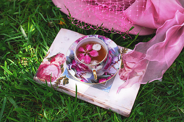 Pink apple tree petals in flower tea in cup on book with hat in grass