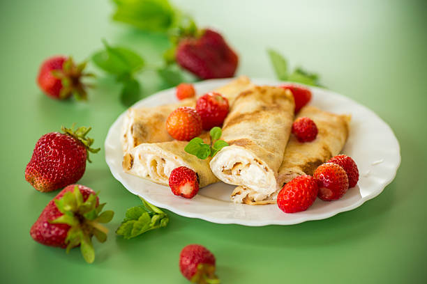 White round plate with crepe pancakes, cut strawberries and vanilla sauce over everything on a white table top, flat lay