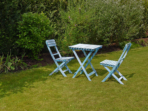 Two blue coloured wooden chairs and a table standing in green grass garden fringed by plants  and bushes. An idyllic place to relax in summer.