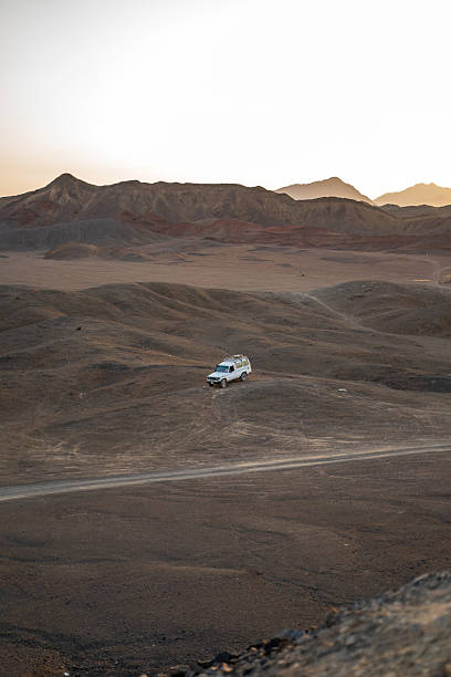istock A white SUV parked amidst a vast and rugged desert terrain, surrounded by distant hills under a golden sunset sky 2219831314