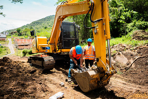 Construction workers examining the bucket of an excavator at an active construction site surrounded by dirt and greenery