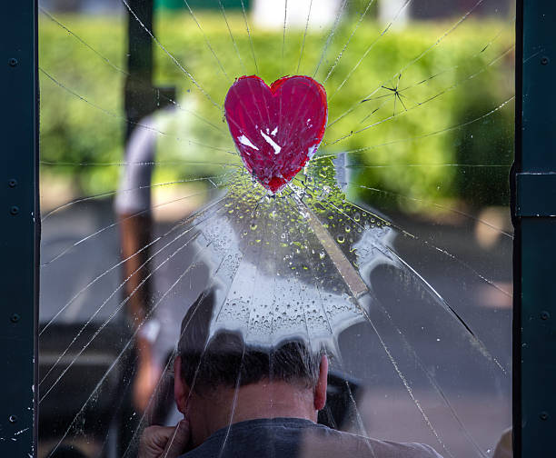 A cracked glass window with a red heart sticker in the center, showing a blurred figure sitting behind it. The background features greenery and hints of urban life.