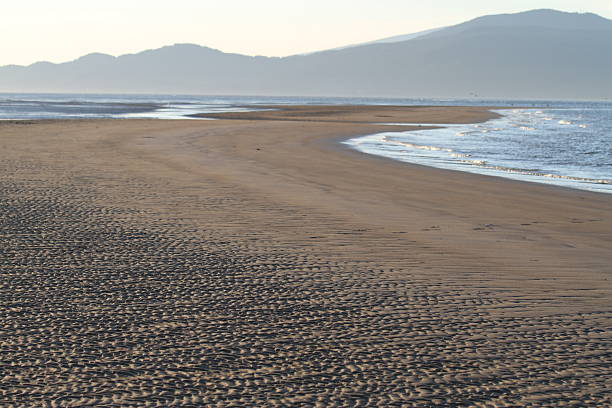 Golden sunlight bathes the expansive sandy shores of Spanish Banks as the sun dips below the horizon, casting warm hues of orange, pink, and purple across the tranquil waters of English Bay. Gentle waves lap the shoreline while silhouettes of distant sailboats and beachgoers add depth to the serene coastal scene. The backdrop of Vancouver’s city skyline and lush North Shore mountains creates a stunning contrast, blending natural beauty with urban charm. This peaceful sunset moment captures the essence of the Pacific Northwest’s scenic coastal landscape, inviting viewers to experience calm, reflection, and the quiet end of a summer day.