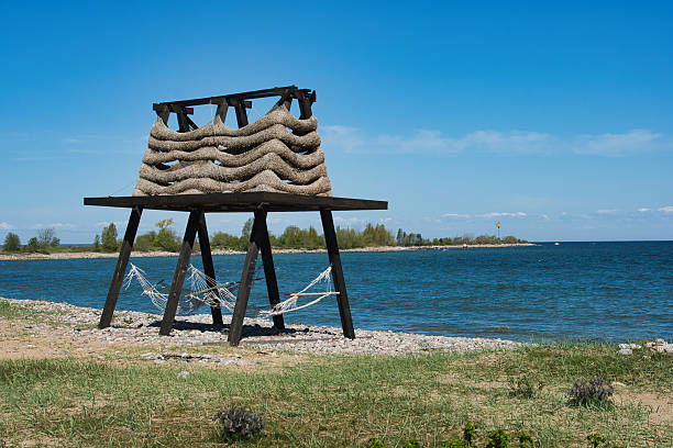 A distinctive wooden hammock stands on a rocky beach in Estonia on the Baltic Sea. The bright blue sky and calm sea create a serene summer backdrop, while the handmade design of the hammocks and the sculptural top evoke a blend of rustic charm and modern creativity. The peaceful coastline, scattered with stones and framed by distant trees, emphasizes relaxation and coastal design harmony.