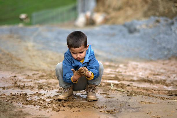 A young boy stands in an open agricultural field on a sunny day, wearing a casual sweater and pants. The rural landscape with hills and farmland stretches behind him, symbolizing childhood in the countryside, connection to nature, and life in farming communities. Concept of rural youth, agriculture, countryside lifestyle, future generation of farmers, and traditional living.