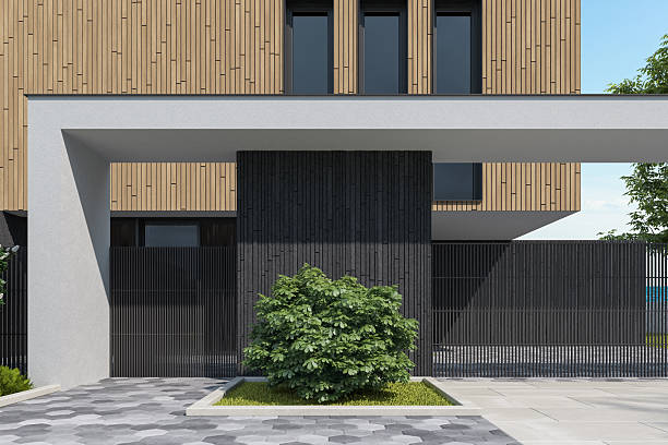 This high-resolution architectural rendering showcases a sleek, modern two-story villa characterized by clean lines, minimalist design, and a harmonious blend of natural and industrial materials. The upper floor facade is elegantly clad in vertical wooden panels, contrasted by dark stone or concrete walls on the ground level. Large vertical windows add rhythm to the structure while allowing abundant natural light to enter the interior. The house features a prominent flat roof, emphasizing its contemporary architectural style.In the foreground, a wide, open driveway with light-gray ceramic or concrete tiles leads to a carport or covered entrance supported by minimalist white beams. A landscaped garden with manicured shrubs, a tall deciduous tree, and a row of evergreen conifers offers a natural touch to the clean geometry of the home. The side pathway includes hexagonal patterned paving stones bordered by grass strips, enhancing the overall visual symmetry and elegance. Perfect for use in real estate promotions, architectural portfolios, construction materials marketing, villa design showcases, and landscaping presentations.