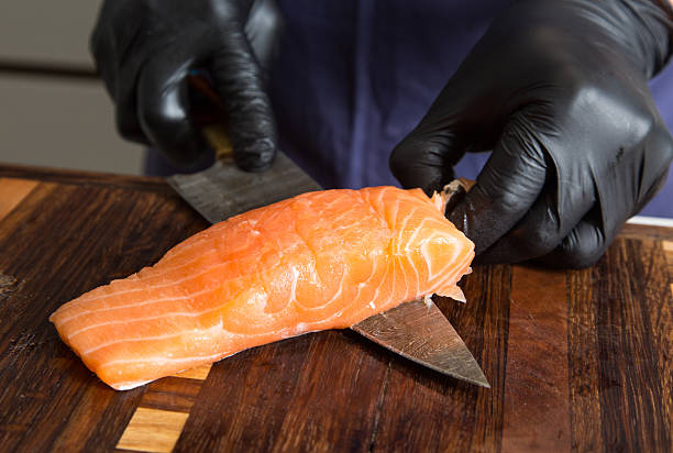 Raw salmon being handled by two hands with black gloves, preparing and cutting with a kitchen knife on a cutting board