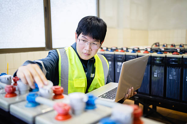 istock Technician wearing safety vest inspects backup power system battery while using laptop to ensure proper maintenance and reliable energy supply in industrial setting 2219874419