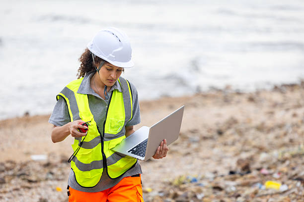 istock Environmental Scientist Conducting Beach Cleanup Survey, Female Engineer Using Laptop for Coastal Area Assessment, Worker in Safety Vest and Hard Hat Documenting Outdoor Site 2219893018