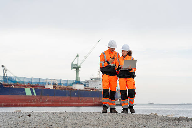 Two engineers wearing high-visibility safety gear and hard hats review plans on a laptop at a shipyard, with a large cargo ship and cranes in the background during sunset.