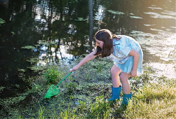 Two young sisters, 10 and 5 years old, are spending a delightful sunny day in a local park. Equipped with a butterfly net and a jar, they enjoy catching and observing butterflies, creating a fun and educational outdoor activity. The green grass and serene river in the background complement their joyful experience.