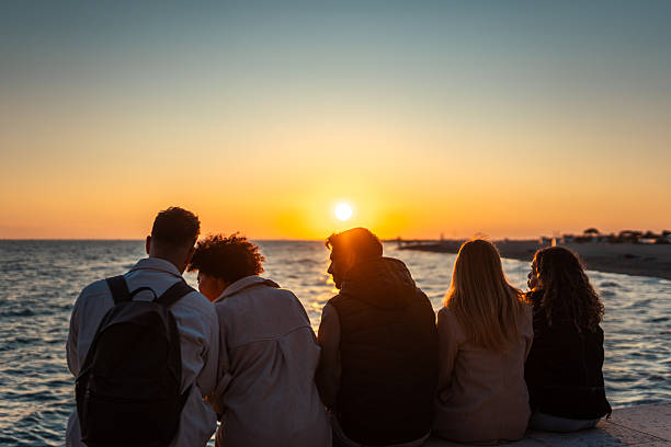 A diverse group of friends sits by the ocean, enjoying a serene sunset. They wear casual clothing, creating a relaxed and peaceful atmosphere.
