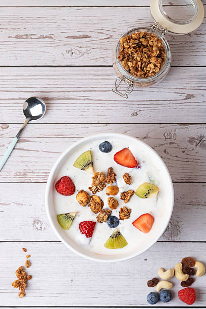 White bowl with kefir, granola, strawberry, kiwi, raspberry, and blueberry, next to a jar of granola and metal spoon. Rustic look, vertical top view. Copy space. Concept of healthy eating and modern lifestyle