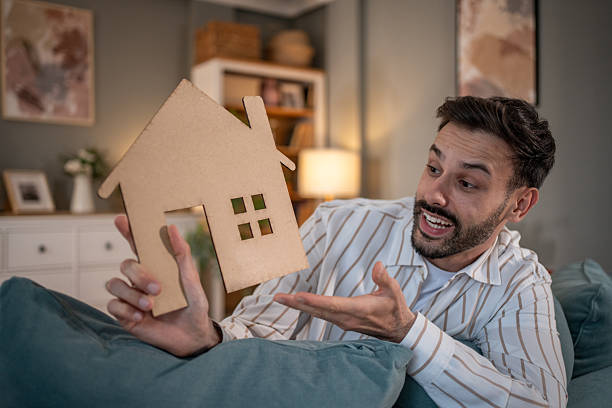 istock Excited man showing a small wooden house while lying on sofa 2219915778
