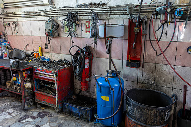Grungy and cluttered automotive workshop interior with various tools, wires, hoses, and equipment mounted on a dirty tiled wall. A red tool chest, fire extinguisher, oil drum, and hanging cables.