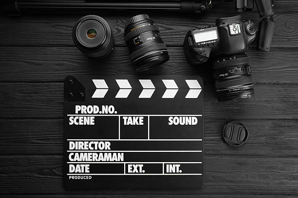 Movie clapper, camera and lens on black wooden table, flat lay