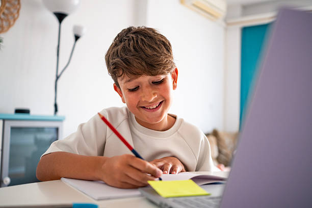 Young boy happily doing homework and writing notes at home while studying with the laptop