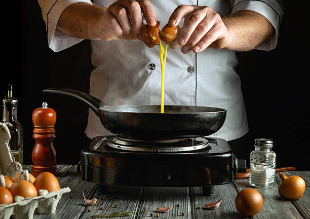 istock A chef in a white coat is cracking eggs into a frying pan placed on a stove. Surrounding the chef are fresh eggs and various kitchen utensils, creating an engaging culinary atmosphere 2219934516