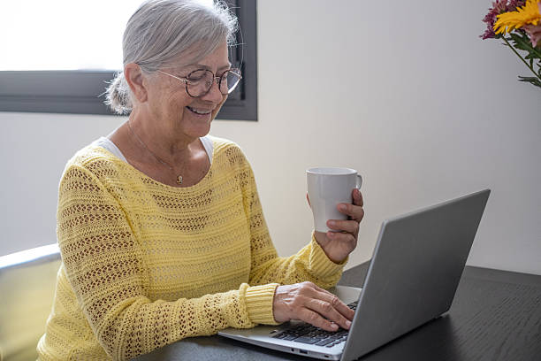 istock Smiling senior grey haired woman sitting at home table using laptop enjoying a coffee cup while browsing on internet 2219947706