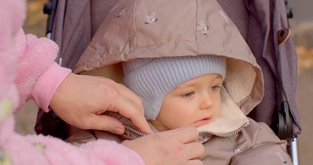 A cheerful toddler in a stroller is cozy and warm, adorned with a snug jacket and a knitted hat, being gently adjusted and cared for by an attentive adult amidst a beautiful park setting