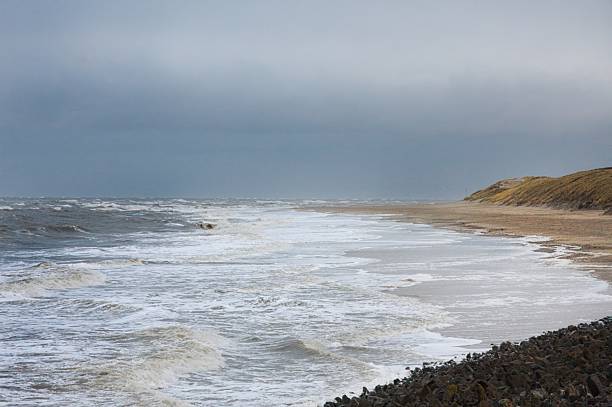 A moody coastal scene showing waves gently breaking onto a broad sandy beach under an overcast sky, with low green hills in the background.