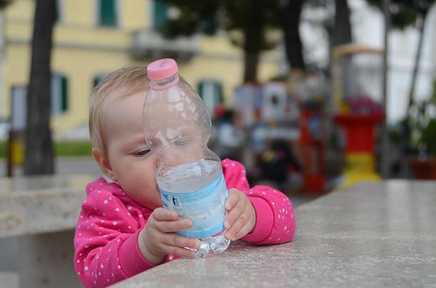 Baby gripping a plastic water bottle with curiosity while sitting at an outdoor table. The child is wearing a pink outfit and focusing intently on the bottle in a playful manner