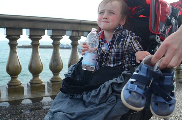 Young child sitting in a stroller by a seaside railing, holding a water bottle while showing a thoughtful expression. An adult hand is holding a pair of blue shoes nearby.