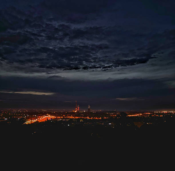 A dramatic view of a cityscape at night, illuminated by glowing streetlights and industrial lights in the distance. Dark, textured storm clouds stretch across the sky, adding depth and contrast to the urban lights below. This image conveys a sense of mystery and quiet energy, ideal for themes of night, city life, weather, and industrial landscapes.
