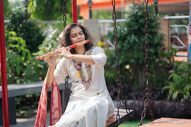 A barefoot woman in traditional attire plays the flute while seated on a swing in a green outdoor courtyard. Her floral embroidery and dupatta add vibrant details to a calm, soulful moment that captures the beauty of Indian culture and mindfulness.