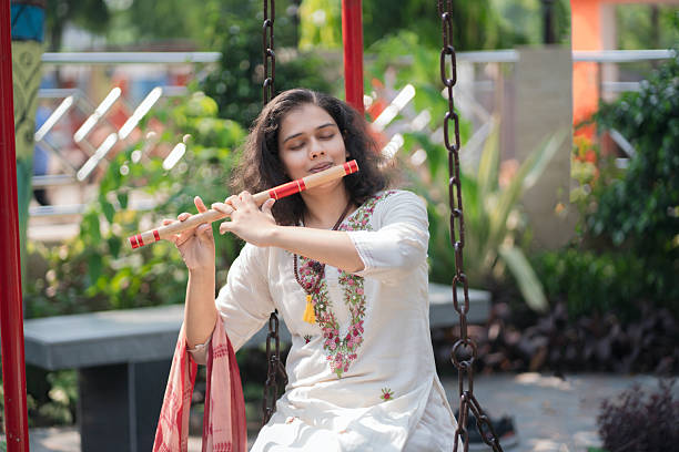 A serene young woman in a traditional embroidered white kurta plays the flute while sitting barefoot on a wooden swing in a lush green garden. Dressed in ethnic Indian attire with a pink dupatta draped over the swing, she appears calm and immersed in her music. The background features vibrant colors, greenery, and outdoor decor, evoking a peaceful and artistic mood.