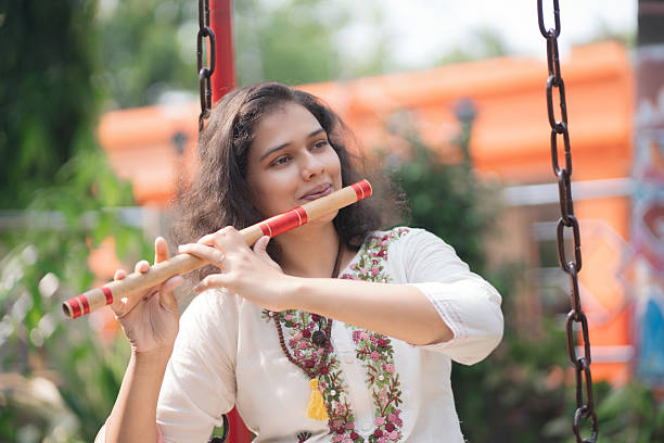 A serene young woman in a traditional embroidered white kurta plays the flute while sitting barefoot on a wooden swing in a lush green garden. Dressed in ethnic Indian attire with a pink dupatta draped over the swing, she appears calm and immersed in her music. The background features vibrant colors, greenery, and outdoor decor, evoking a peaceful and artistic mood.