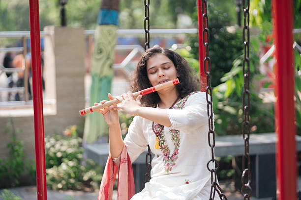A barefoot woman in traditional attire plays the flute while seated on a swing in a green outdoor courtyard. Her floral embroidery and dupatta add vibrant details to a calm, soulful moment that captures the beauty of Indian culture and mindfulness.