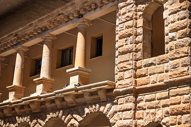 Part of a historic Mediterranean building facade with stone arches, Doric columns, and textured walls. Sunlight casts shadows, enhancing the warm tones and architectural detail.