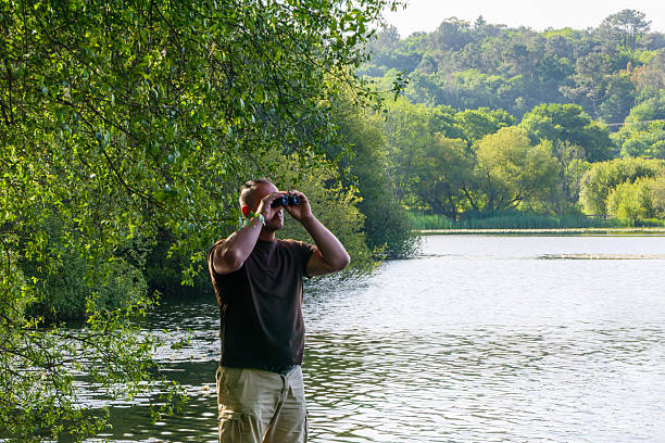 A man in a dark t-shirt and beige shorts watches the scenery through binoculars from the lakeshore, surrounded by lush greenery. The peaceful environment is ideal for birdwatching.