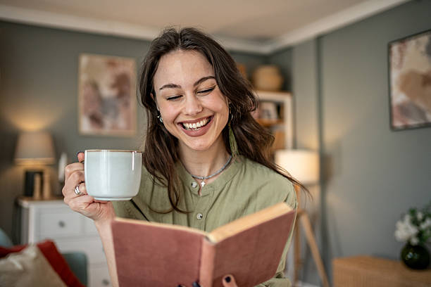 istock Young woman laughing while reading a book and drinking coffee at home 2219998974