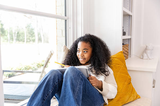 Smiling girl sits on window seat writing in notebook. Casual, cozy, and happy.