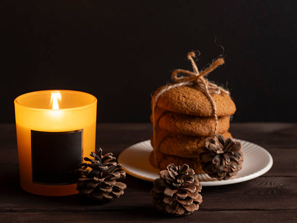 A cozy still life featuring a stack of homemade cookies tied with twine, next to a burning scented candle and pinecones on a dark wooden surface. Warm and rustic holiday or autumn atmosphere