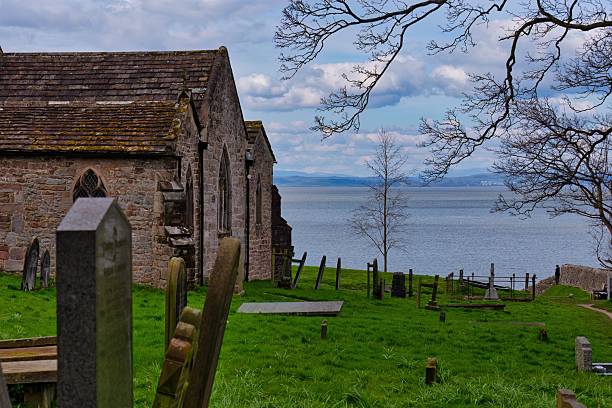 An old stone building sits beside a green graveyard overlooking a body of water under a cloudy sky.