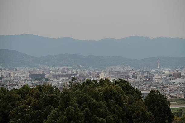 The skyline of Kyoto stretches beneath a soft sky, with the iconic Kyoto Tower rising prominently above the cityscape. Traditional low-rise buildings blend with modern structures, capturing the balance between history and progress. In the distance, hazy mountains form a natural border, while the tower’s observation deck gleams in the light, offering panoramic views of the ancient capital reborn in the present.