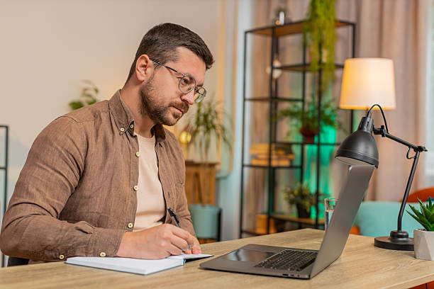 istock Young freelancer writing down notes while attending online meeting on laptop at home office desk 2220025156