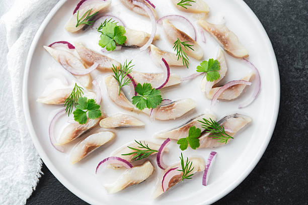 istock Marinated herring slices with red onion and herbs on plate with napkin on dark background 2220025282