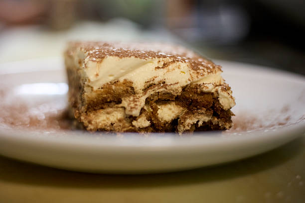 A close-up of a single slice of tiramisu on a white plate, its creamy layers dusted with cocoa and softly lit.