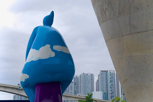 Incheon, South Korea - May 19th 2025: The whimsical back of the Cycling Cloud Man sculpture faces a cluster of high-rise apartments and the Ara Waterway's monorail bridge, beneath the towering Sicheon Bridge in Sicheon Waterside Park.