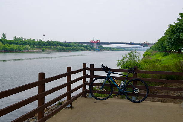 Incheon, South Korea - May 19th 2025: A road bike rests against a wooden fence beside the Ara Waterway as Baekun Bridge stretches across the canal in the distance, framed by green riverside trails.
