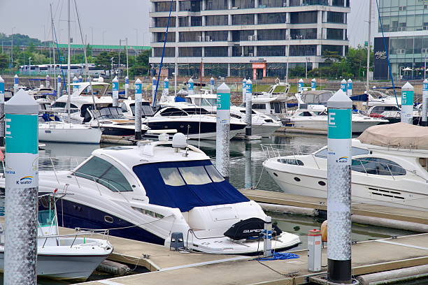 Incheon, South Korea - May 19th 2025: A row of yachts, including cabin cruisers and powerboats, are docked at Pier E of Ara Marina, with residential and commercial buildings lining the waterfront in the background.