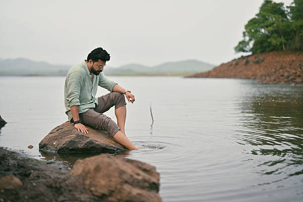 A man sits alone on a rock at the edge of a calm lake with his feet in the water. He appears lost in thought as he looks into the water. Gentle hills and greenery surround the peaceful setting, creating a quiet atmosphere ideal for reflection.