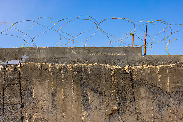 A weathered concrete wall topped with sharp barbed wire under a cloudless blue sky. The rough texture of the wall and the menacing spirals of the wire create a stark and secure boundary.