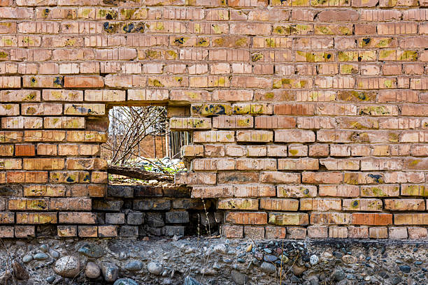 A section of an old brick wall with rectangular hole revealing a glimpse of the outdoors with some branches. The weathered texture of the wall displays cracks and damage, with a mix of brown and yellowish tones.